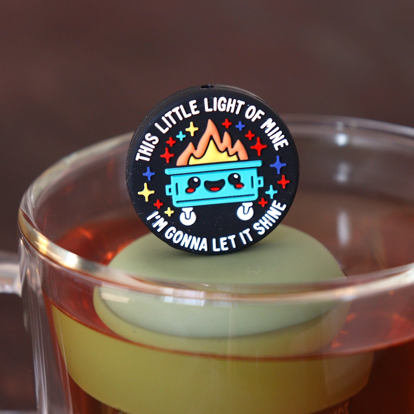 black circle that reads "this little light of mine I'm gonna let it shine" surrounding a smiling dumpster tea infuser for loose leaf tea  with a fine mesh stainless steel tea infuser that reads FLOAT2INFUSE on a green base floating in a glass tea mug