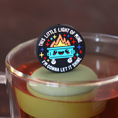 black circle that reads "this little light of mine I'm gonna let it shine" surrounding a smiling dumpster tea infuser for loose leaf tea  with a fine mesh stainless steel tea infuser that reads FLOAT2INFUSE on a green base floating in a glass tea mug