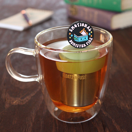 black circle with text that reads "emotional dumpster fire" and an image of a smiling dumpster fire tea infuser for loose leaf tea with a fine mesh stainless steel tea infuser that reads FLOAT2INFUSE  on a green base floating in a glass tea mug with pen on notebook and books in the background