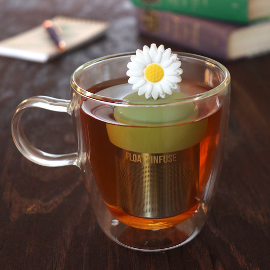 giant white daisy tea infuser for loose leaf tea with a fine mesh stainless steel tea infuser that reads FLOAT2INFUSE on a green base  floating in a glass tea mug with pen on notebook and loose leaf tea and books in the background