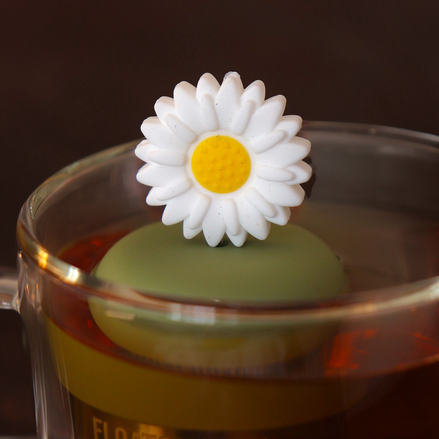 giant white daisy tea infuser for loose leaf tea  with a fine mesh stainless steel tea infuser that reads FLOAT2INFUSE on a green base floating in a glass tea mug 