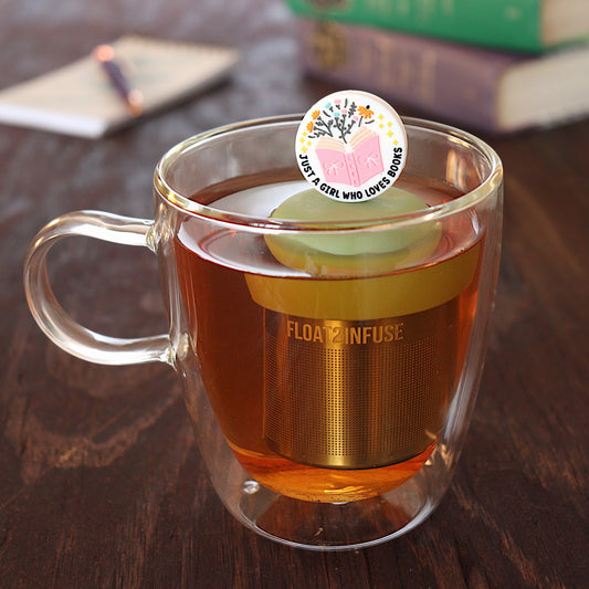 white circle that reads "just a girl who loves books" with stars and flowers coming out of a pink book tea infuser for loose leaf tea with a fine mesh stainless steel tea infuser that reads FLOAT2INFUSE on a green base  floating in a glass tea mug with pen on notebook and loose leaf tea and books in the background
