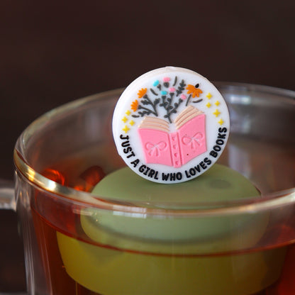 white circle that reads "just a girl who loves books" with stars and flowers coming out of a pink book tea infuser for loose leaf tea  with a fine mesh stainless steel tea infuser that reads FLOAT2INFUSE on a green base floating in a glass tea mug