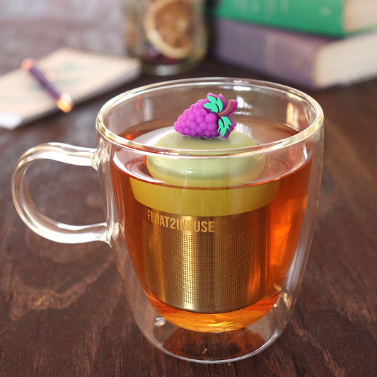 purple grapes tea infuser for loose leaf tea with a fine mesh stainless steel tea infuser that reads FLOAT2INFUSE on a green base  floating in a glass tea mug with pen on notebook and loose leaf tea and books in the background
