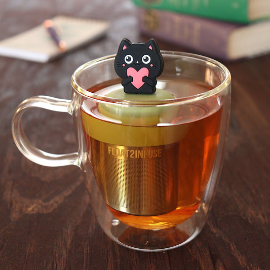 black cat animal holding pink heart tea infuser for loose leaf tea on a green base with a fine mesh stainless steel tea infuser that reads FLOAT2INFUSE floating in a glass tea mug with pen on notebook and books in the background