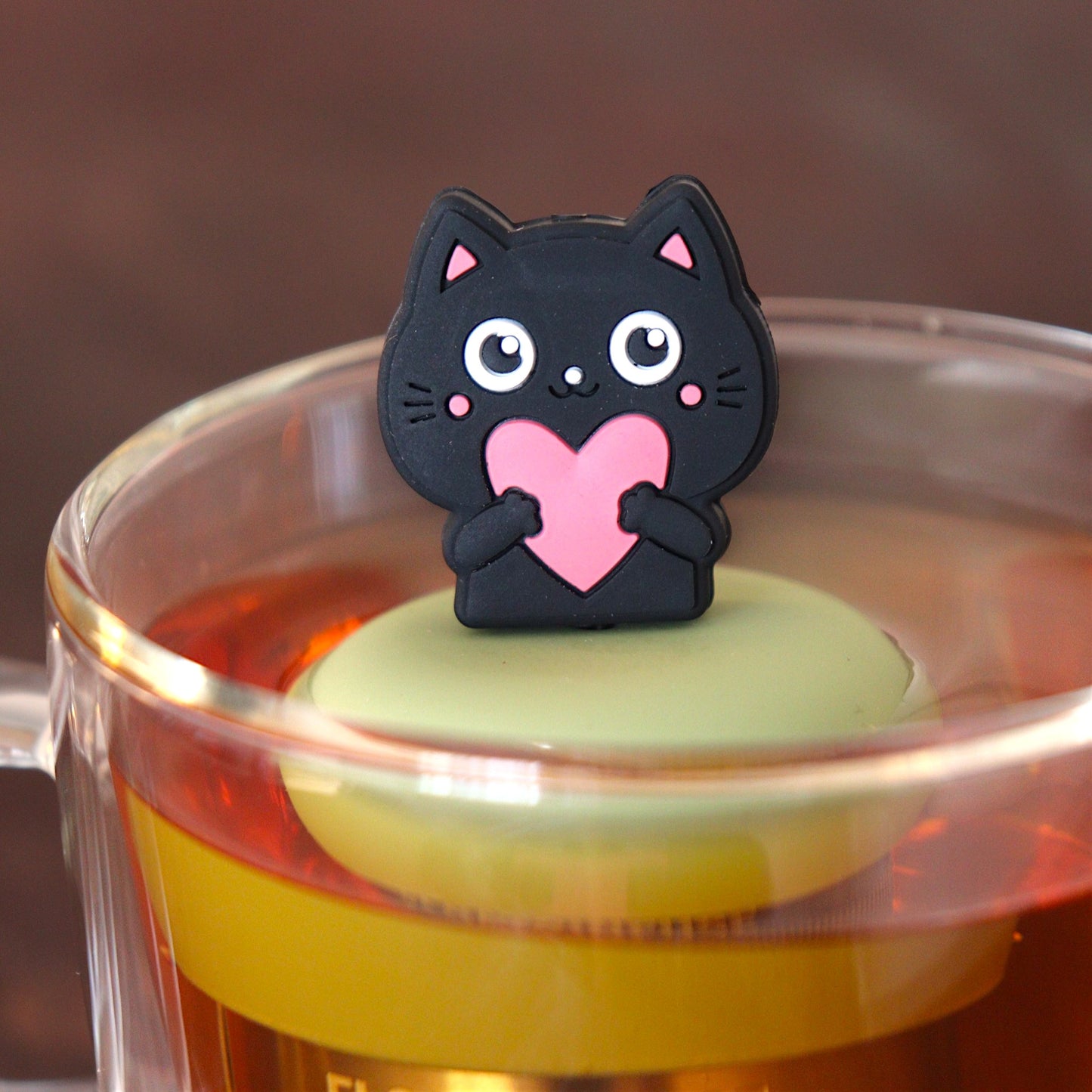 black cat animal holding pink heart tea infuser for loose leaf tea  with a fine mesh stainless steel tea infuser that reads FLOAT2INFUSE on a green base floating in a glass tea mug