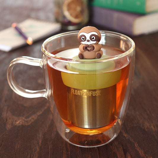 brown sloth animal creating a heart tea infuser for loose leaf tea  with a fine mesh stainless steel tea infuser that reads FLOAT2INFUSE on a green base floating in a glass tea mug with pen on notebook and loose leaf tea and books in the background