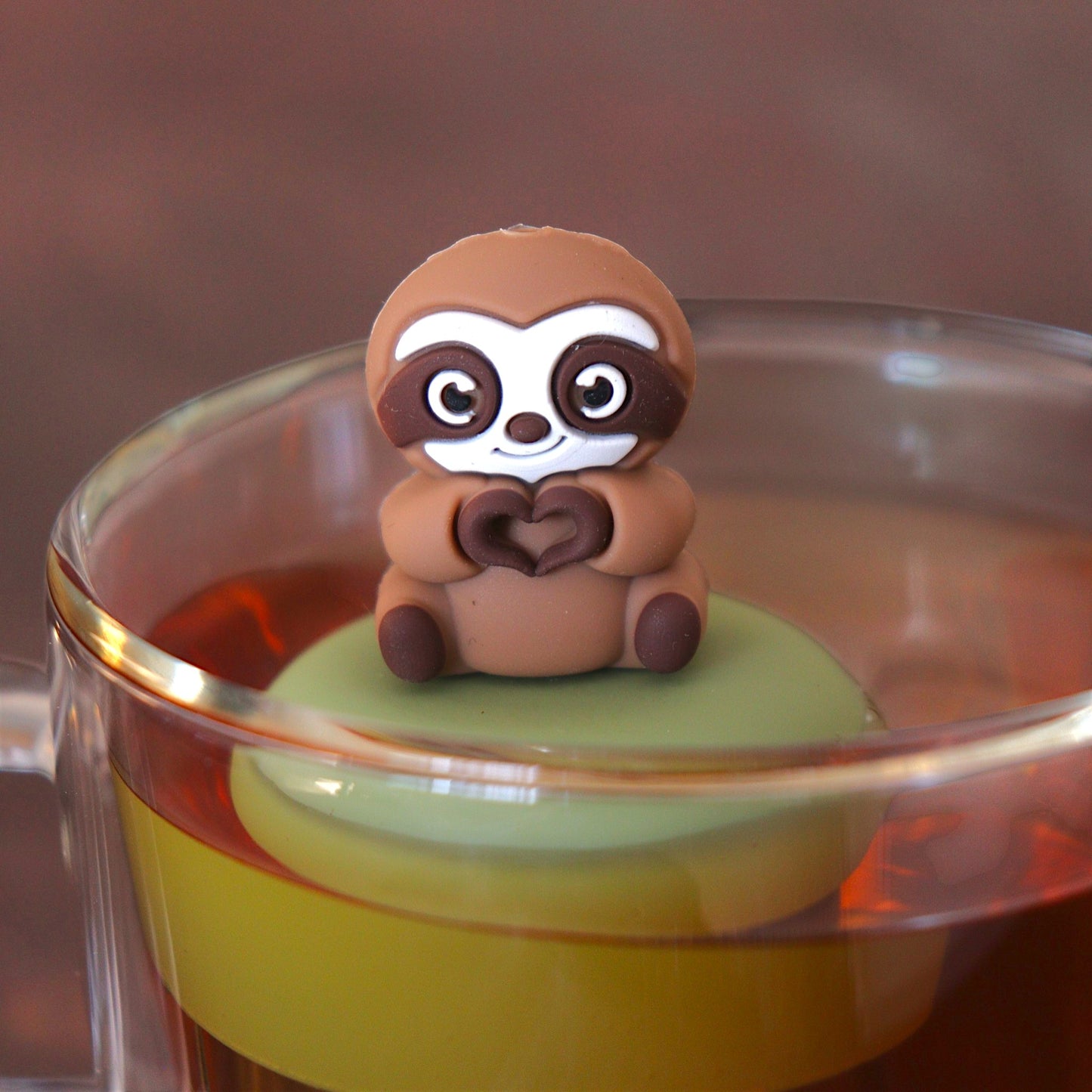brown sloth animal creating a heart tea infuser for loose leaf tea with a fine mesh stainless steel tea infuser that reads FLOAT2INFUSE  on a green base floating in a glass tea mug