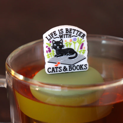 text on white background that reads "life is better with cats & books" with flowers and black cat sitting on an open book tea infuser for loose leaf tea  with a fine mesh stainless steel tea infuser that reads FLOAT2INFUSE on a green base floating in a glass tea mug
