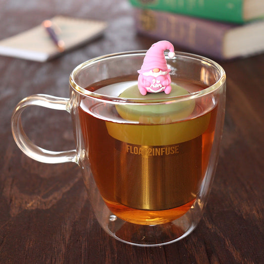 pink gnome holding a heart that reads "i love mom" tea infuser for loose leaf tea  with a fine mesh stainless steel tea infuser that reads FLOAT2INFUSE on a green base floating in a glass tea mug with pen on notebook and loose leaf tea and books in the background
