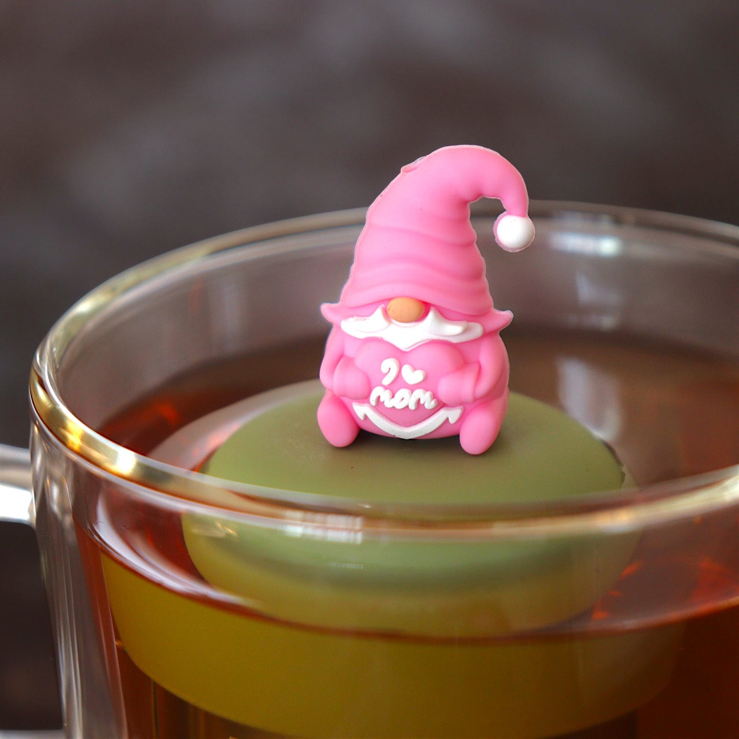 pink gnome holding a heart that reads "i love mom" tea infuser for loose leaf tea  with a fine mesh stainless steel tea infuser that reads FLOAT2INFUSE on a green base floating in a glass tea mug
