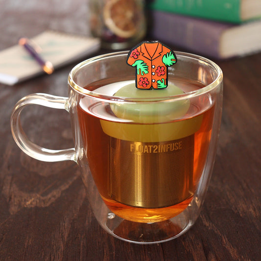 orange hawaiian shirt tea infuser for loose leaf tea on a green base with a fine mesh stainless steel tea infuser that reads FLOAT2INFUSE floating in a glass tea mug with pen on notebook and loose leaf tea and books in the background