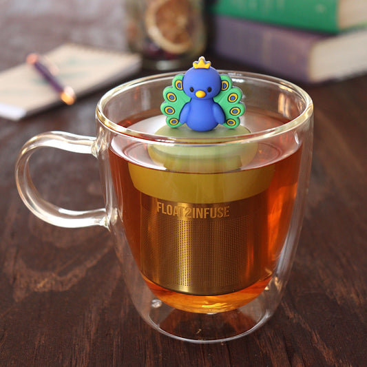blue and green and yellow peacock animal tea infuser for loose leaf tea  with a fine mesh stainless steel tea infuser that reads FLOAT2INFUSE on a green base floating in a glass tea mug with pen on notebook and loose leaf tea and books in the background