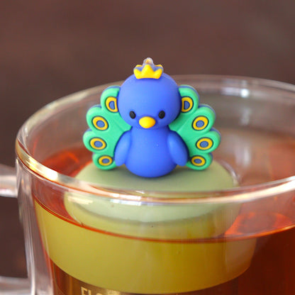blue and green and yellow peacock animal tea infuser for loose leaf tea  with a fine mesh stainless steel tea infuser that reads FLOAT2INFUSE on a green base floating in a glass tea mug