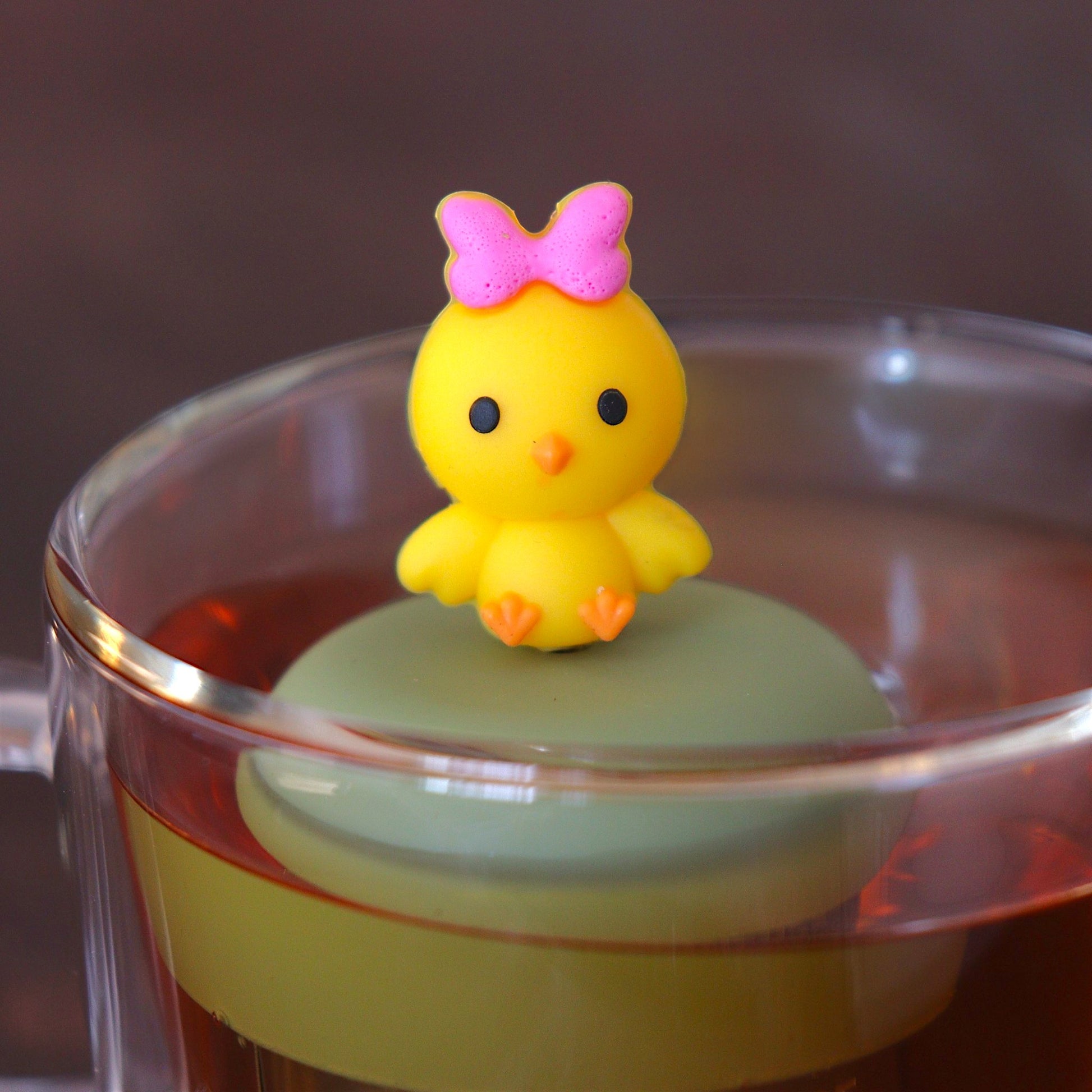 yellow chick with pink bow tea infuser for loose leaf tea  with a fine mesh stainless steel tea infuser that reads FLOAT2INFUSE on a green base floating in a glass tea mug