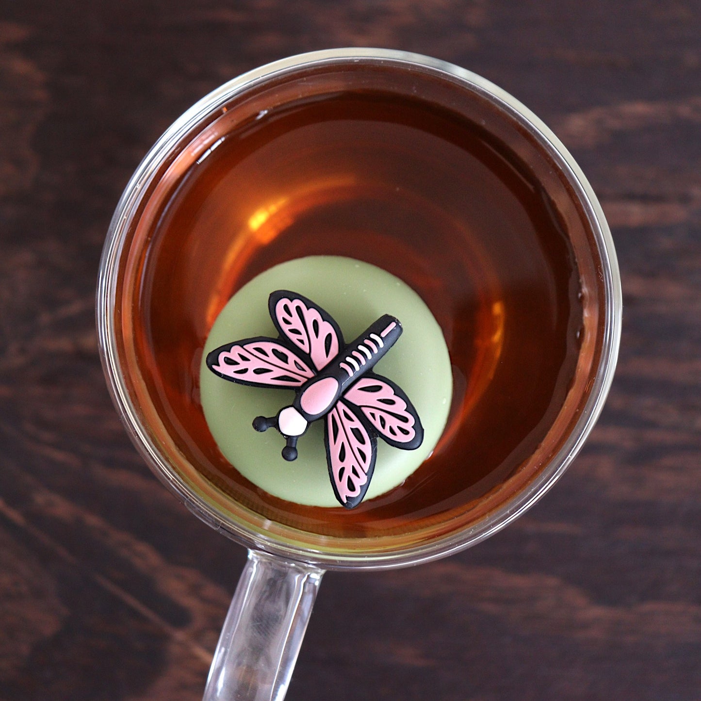 Top view of a pink dragonfly tea infuser for loose leaf tea with a fine mesh stainless steel tea strainer 
that reads FLOAT2INFUSE on a green base floating in a glass tea mug 
