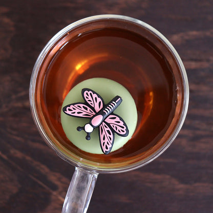 Top view of a pink dragonfly tea infuser for loose leaf tea with a fine mesh stainless steel tea strainer 
that reads FLOAT2INFUSE on a green base floating in a glass tea mug 

