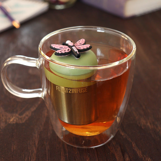 Blue dragonfly tea infuser for loose leaf tea with a fine mesh stainless steel tea strainer 
that reads FLOAT2INFUSE on a green base with a notebook and pen in the background