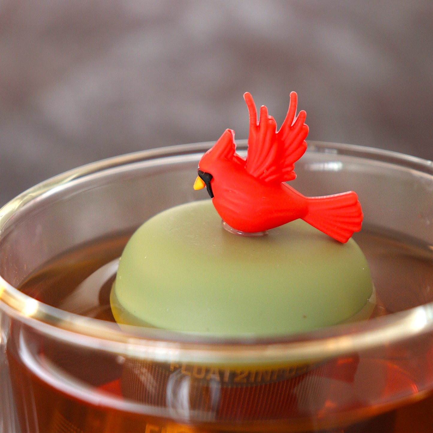 Red cardinal bird tea infuser for loose leaf tea with a fine mesh stainless steel tea strainer that reads FLOAT2INFUSE on a green base floating in a glass tea mug