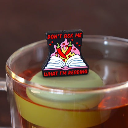 black background with text that reads "don't ask me what I'm reading" with red peppers and flames coming out of an open book tea infuser for loose leaf tea  with a fine mesh stainless steel tea infuser that reads FLOAT2INFUSE on a green base floating in a glass tea mug 