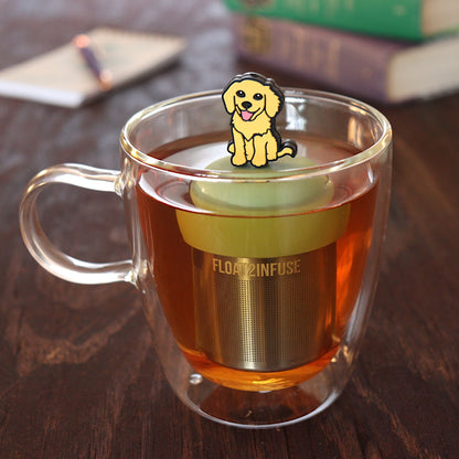 yellow smiling dog animal tea infuser for loose leaf tea on a green base with a fine mesh stainless steel tea infuser that reads FLOAT2INFUSE floating in a glass tea mug with pen on notebook and loose leaf tea and books in the background