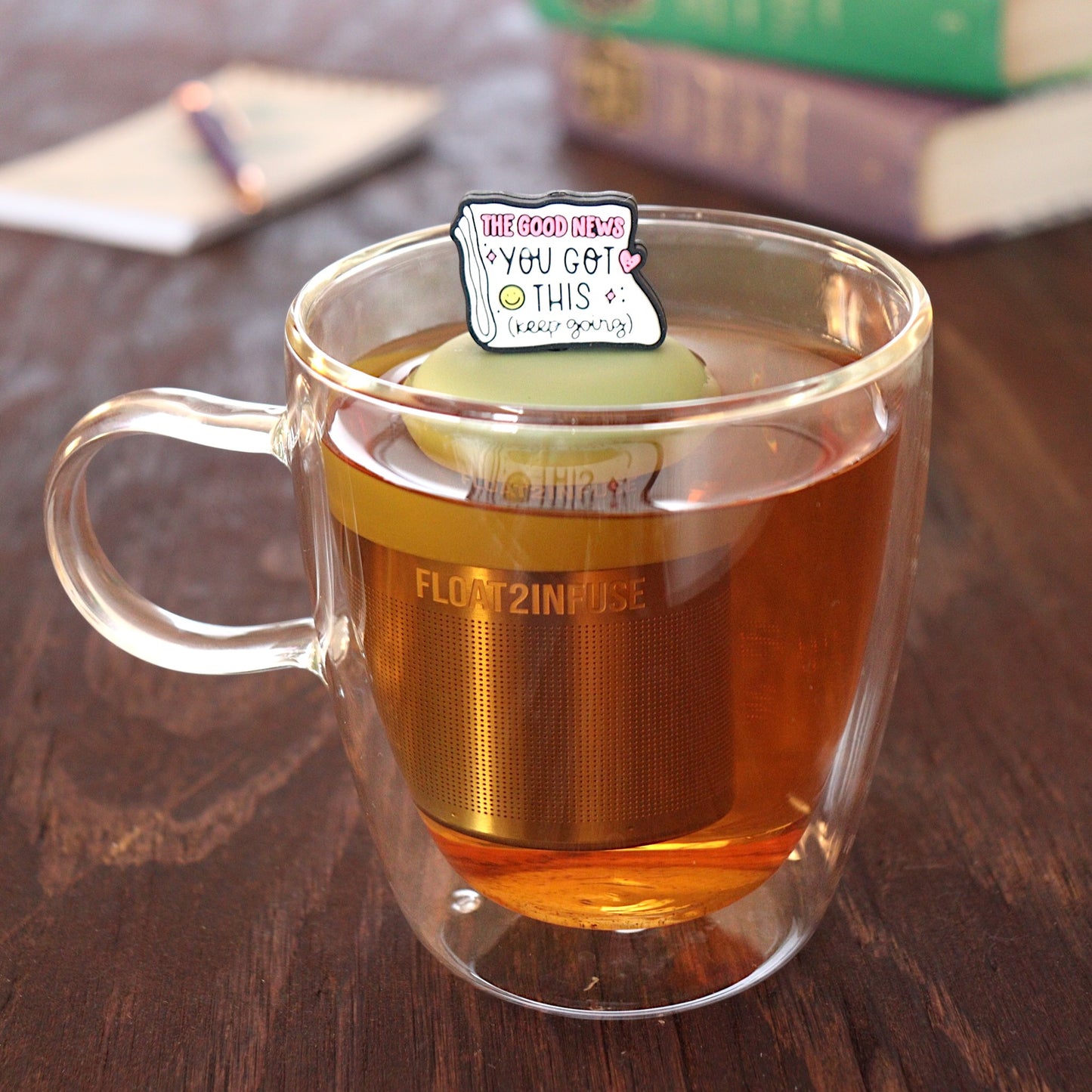 Folded newspaper with text that reads "The good news. You've got this. Keep going" with a pink heart and a yellow smiley face tea infuser for loose leaf tea with a fine mesh stainless steel tea strainer 
that reads FLOAT2INFUSE on a green base floating in a glass tea mug with a pen laying on a notebook and books in the background
 
