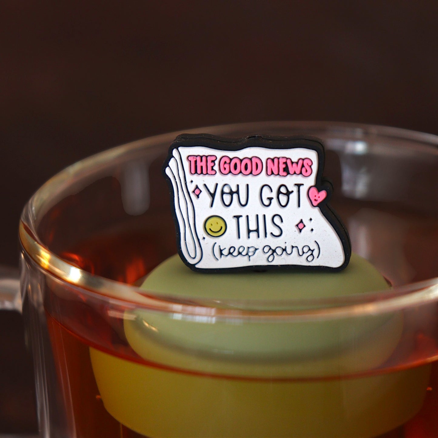 Folded newspaper with text that reads "The good news. You've got this. Keep going" with a pink heart and a yellow smiley face tea infuser for loose leaf tea with a fine mesh stainless steel tea strainer 
that reads FLOAT2INFUSE on a green base floating in a glass tea mug 
 