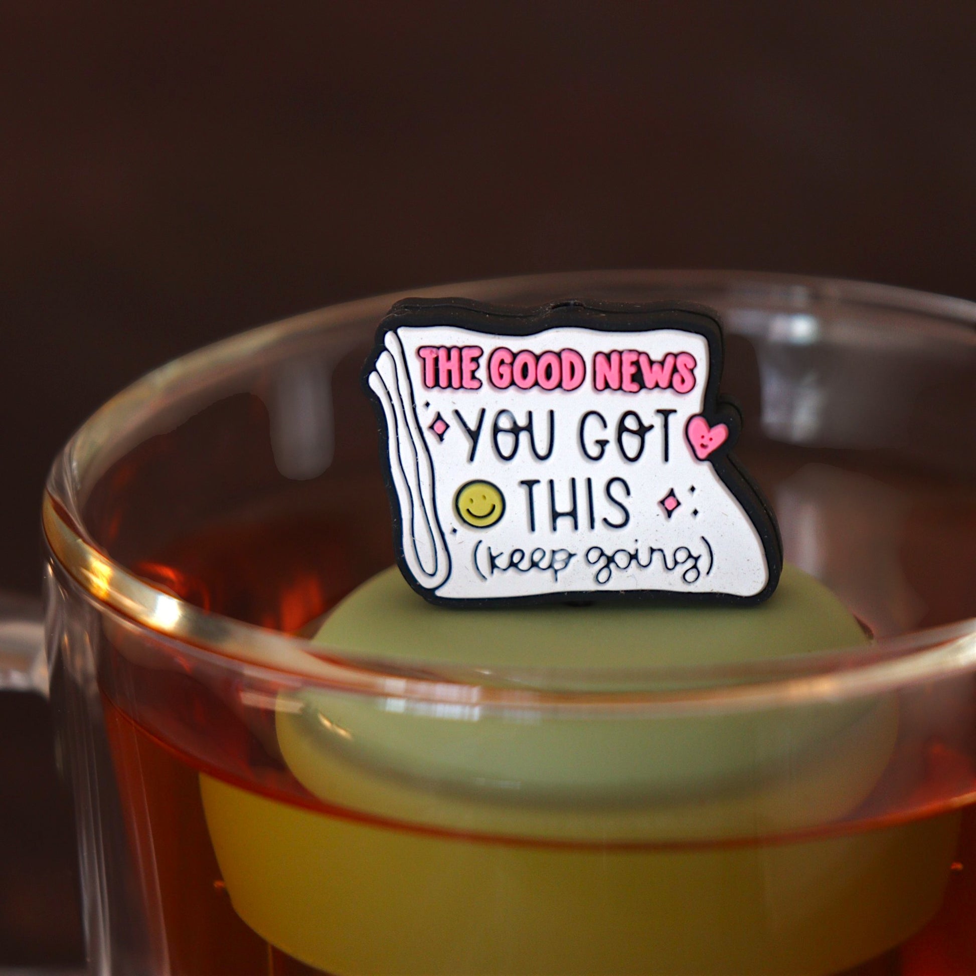 Folded newspaper with text that reads "The good news. You've got this. Keep going" with a pink heart and a yellow smiley face tea infuser for loose leaf tea with a fine mesh stainless steel tea strainer 
that reads FLOAT2INFUSE on a green base floating in a glass tea mug 
 