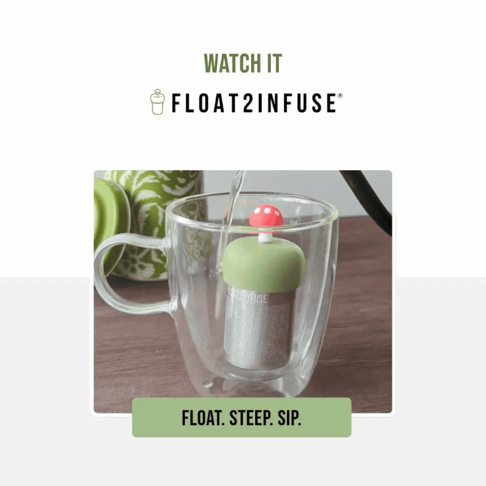 A floating tea infuser in a clear double-walled glass mug, with water being poured into it. The infuser has a light green top, a silver fine mesh stainless steel strainer, and a small red mushroom cap with white dots.