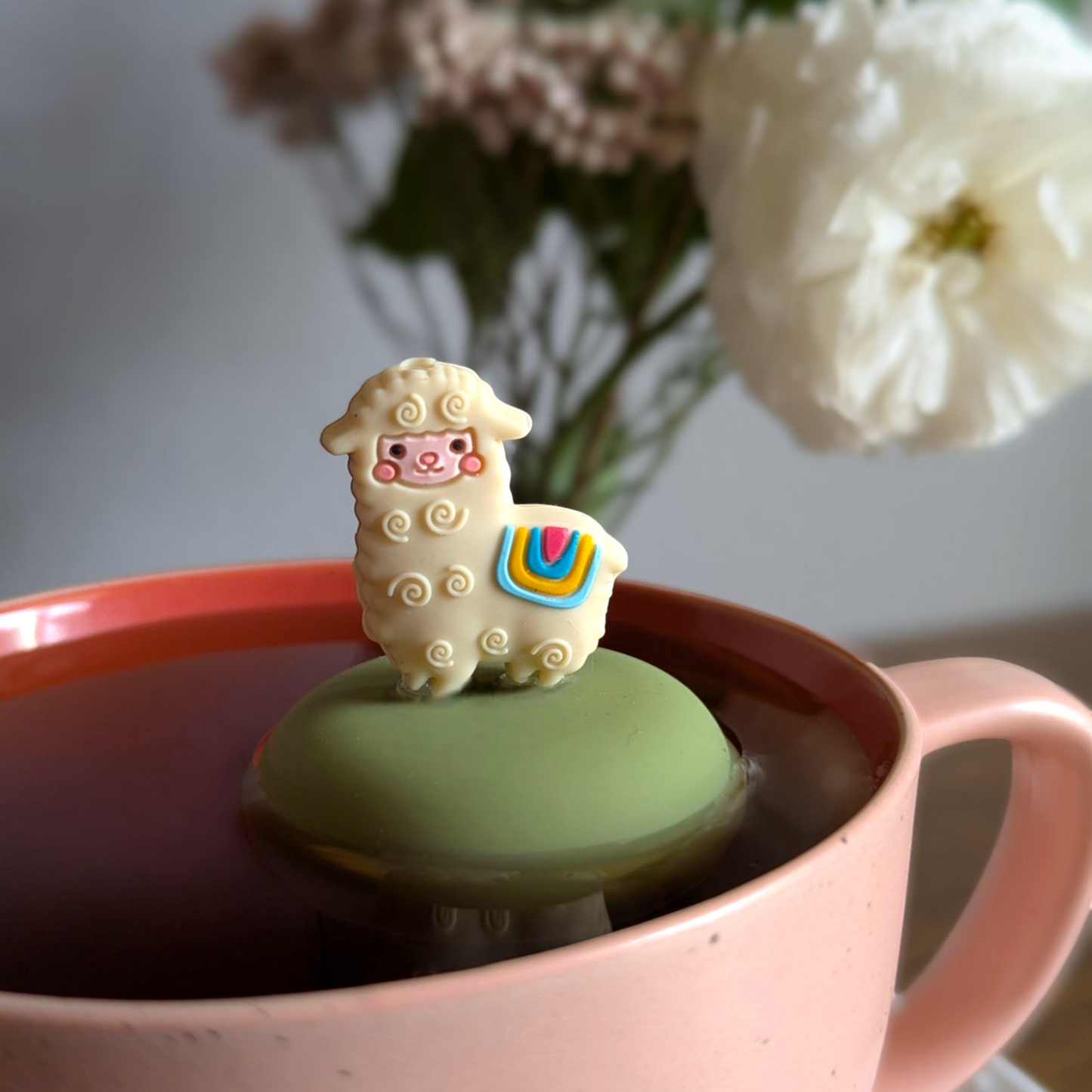 cute alpaca animal tea infuser for loose leaf tea with a fine mesh stainless steel tea strainer that reads FLOAT2INFUSE on a green base floating in a pink tea mug with flowers in the background