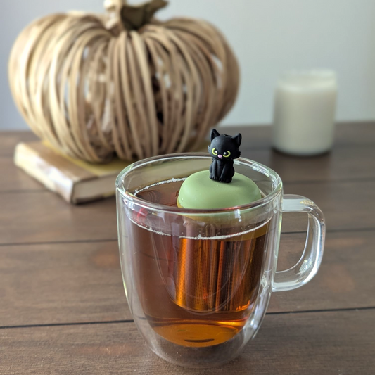 A glass cup with a black cat-shaped floating tea steeper in it, surrounded by a decorative pumpkin and a candle on a wooden surface.