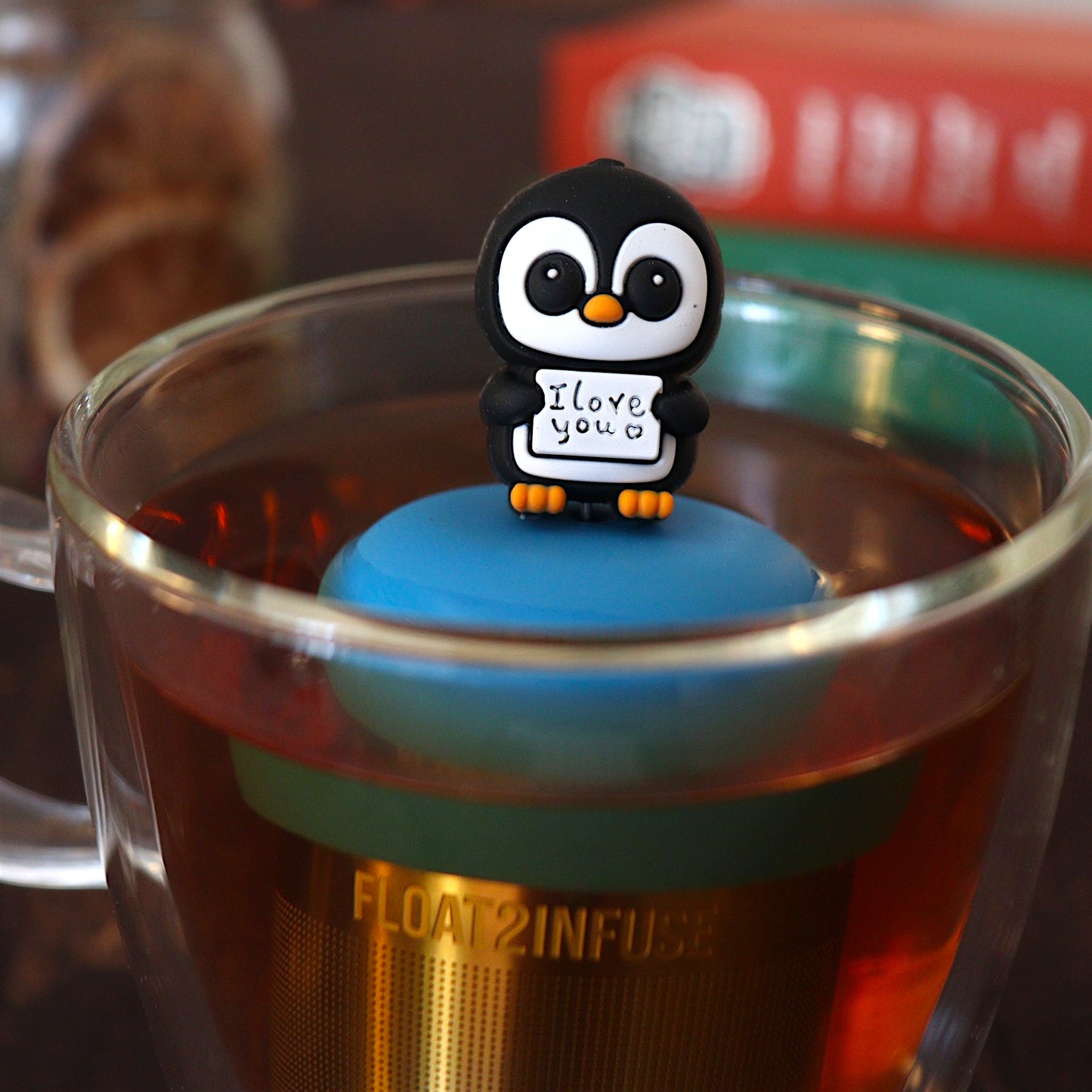 black and white penguin animal holding a sign with text that reads "i love you" tea infuser for loose leaf tea  with a fine mesh stainless steel tea infuser that reads FLOAT2INFUSE on a blue base floating in a glass tea mug with books in the background