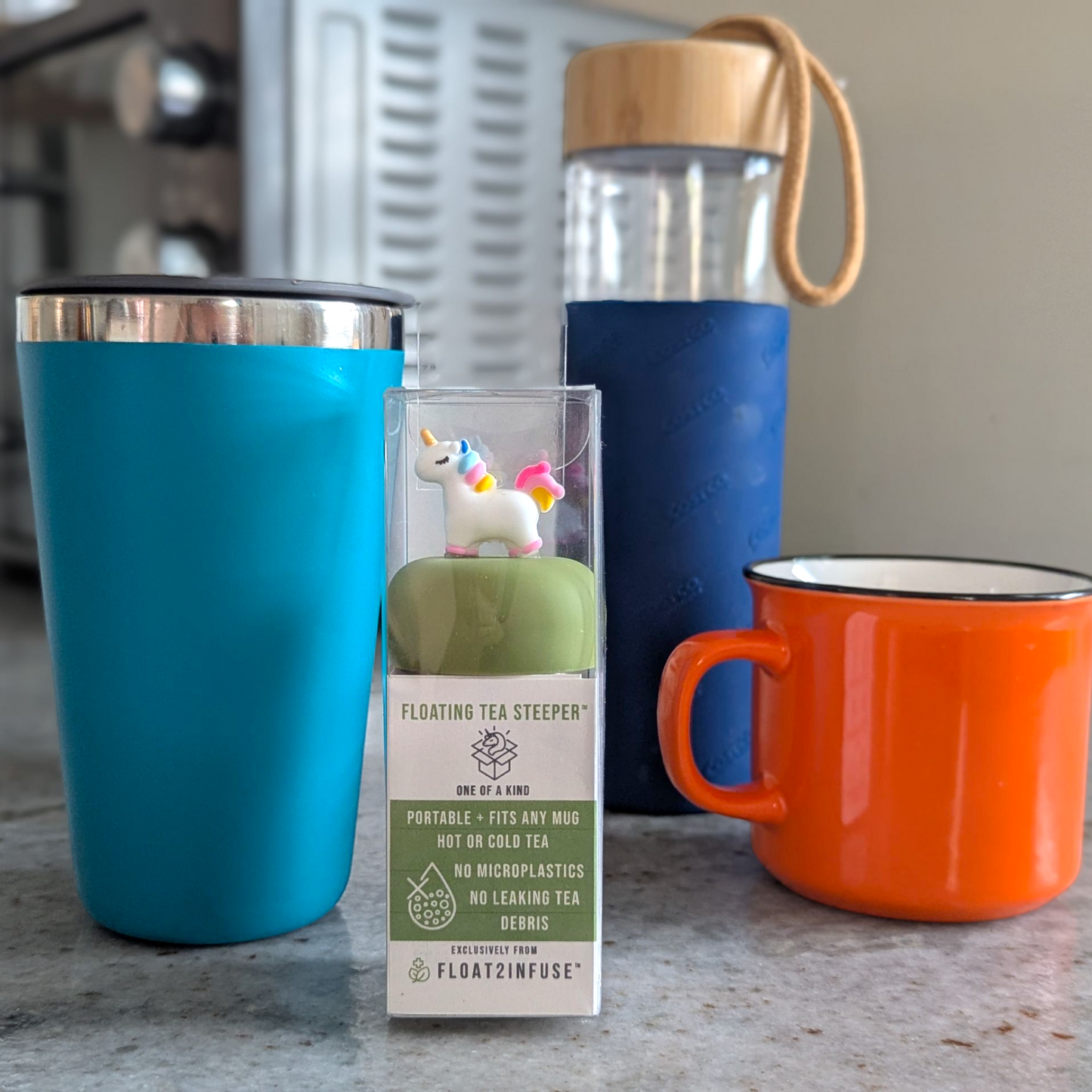 floating unicorn tea infuser in gift box packaging beside a blue tumbler, glass tea tumbler bottle , and orange mug on a kitchen countertop.