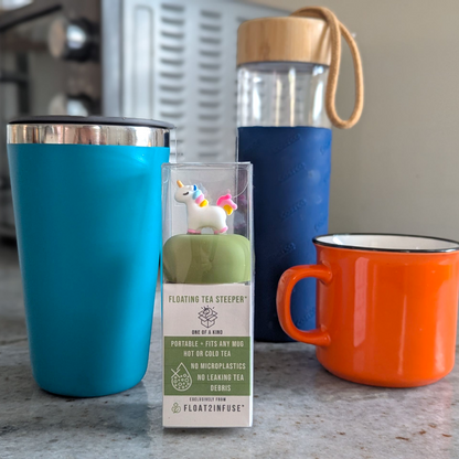 floating unicorn tea infuser in gift box packaging beside a blue tumbler, glass tea tumbler bottle , and orange mug on a kitchen countertop.