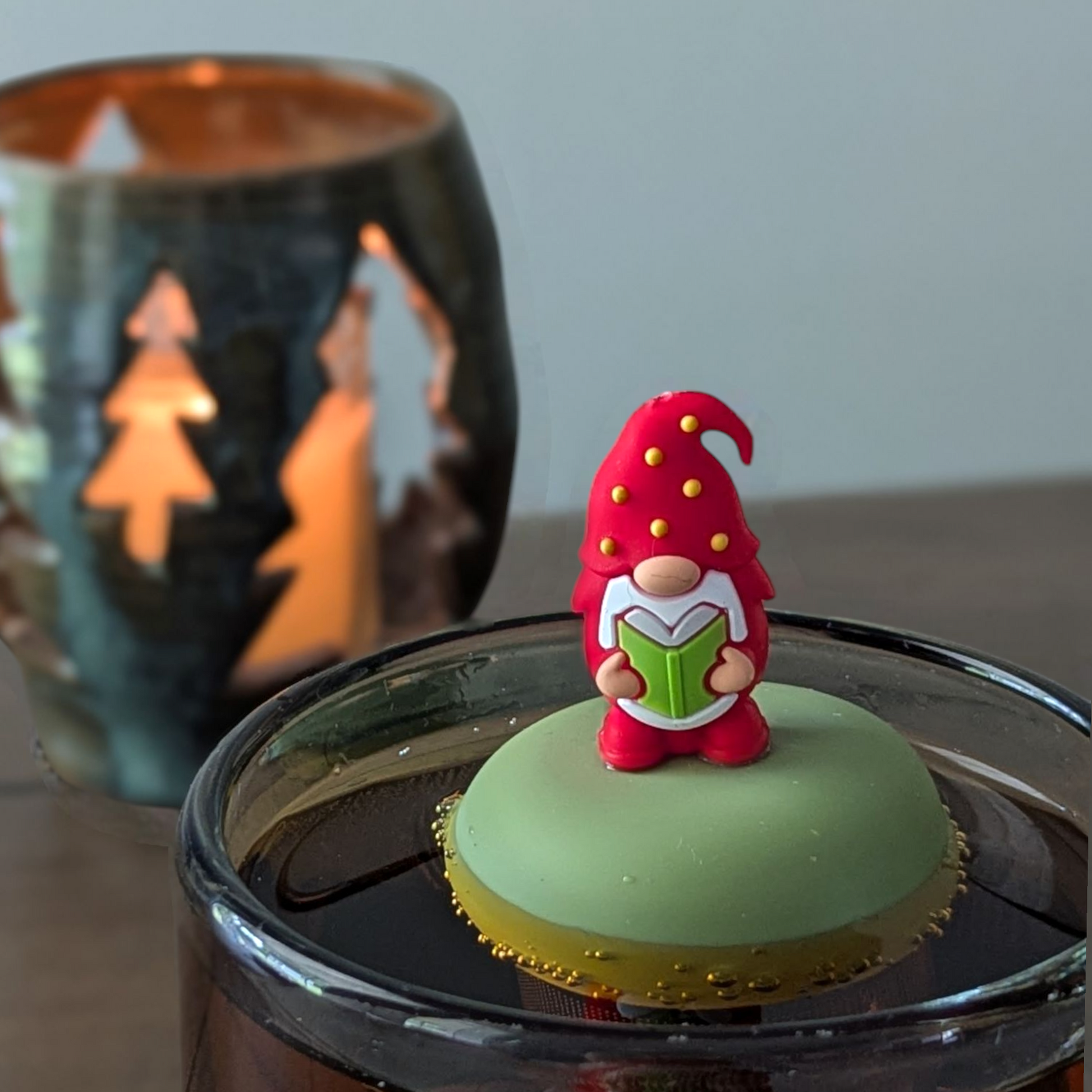 gnome with a long white beard wearing red with yellow polka dots on the hat and holding a green book  tea infuser for loose leaf tea with a fine mesh stainless steel tea strainer that reads FLOAT2INFUSE on a green base  floating in a glass tea mug with a Christmas candle in the background
