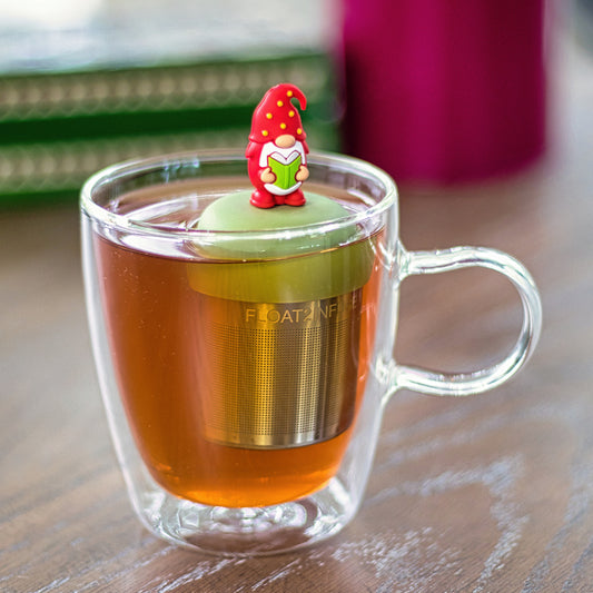 gnome with a long white beard wearing red with yellow polka dots on the hat and holding a green book  tea infuser for loose leaf tea with a fine mesh stainless steel tea strainer that reads FLOAT2INFUSE on a green base  floating in a glass tea mug 
with books in the background