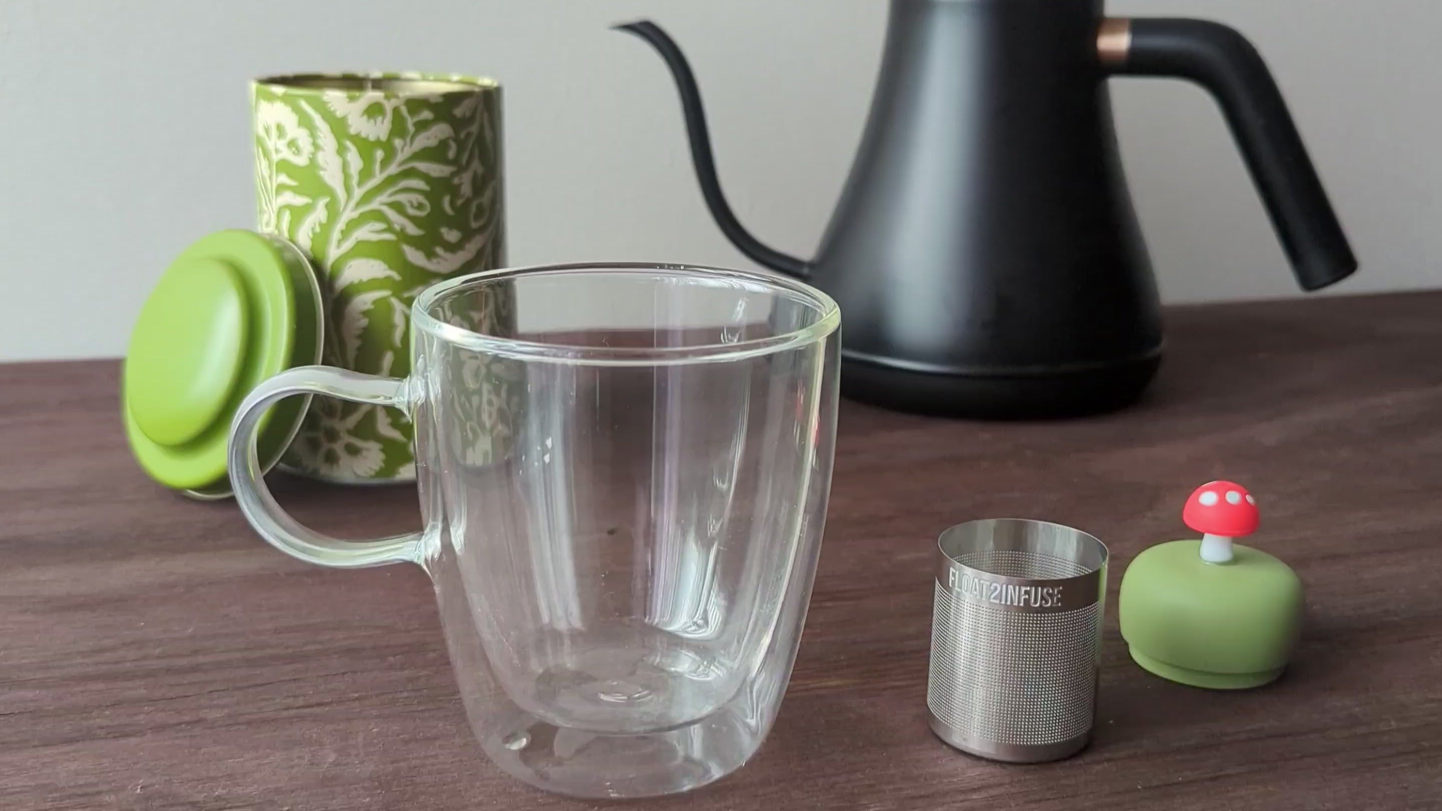 Load video: A step by step video tutorial for how to use a Float2infuse tea infuser. The tea infuser is sitting on a wooden table. A person puts 3 teaspoons of loose tea leaves into the ultra fine mesh stainless steel tea strainer. The green silicone floater of the red mushroom tea infuser is pushed onto the tea basket. The floating tea infuser is placed in a glass mug. hot water is poured into the glass tea cup using a goose neck tea kettle. the floating tea infuser immediately floats and infuses the water to brew hot tea.