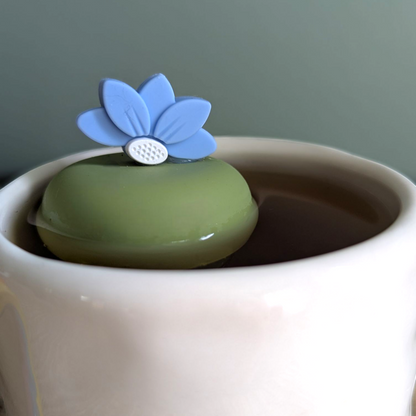 blue lotus flower tea infuser for loose leaf tea  with a fine mesh stainless steel tea infuser that reads FLOAT2INFUSE on a green base floating in a white tea mug 