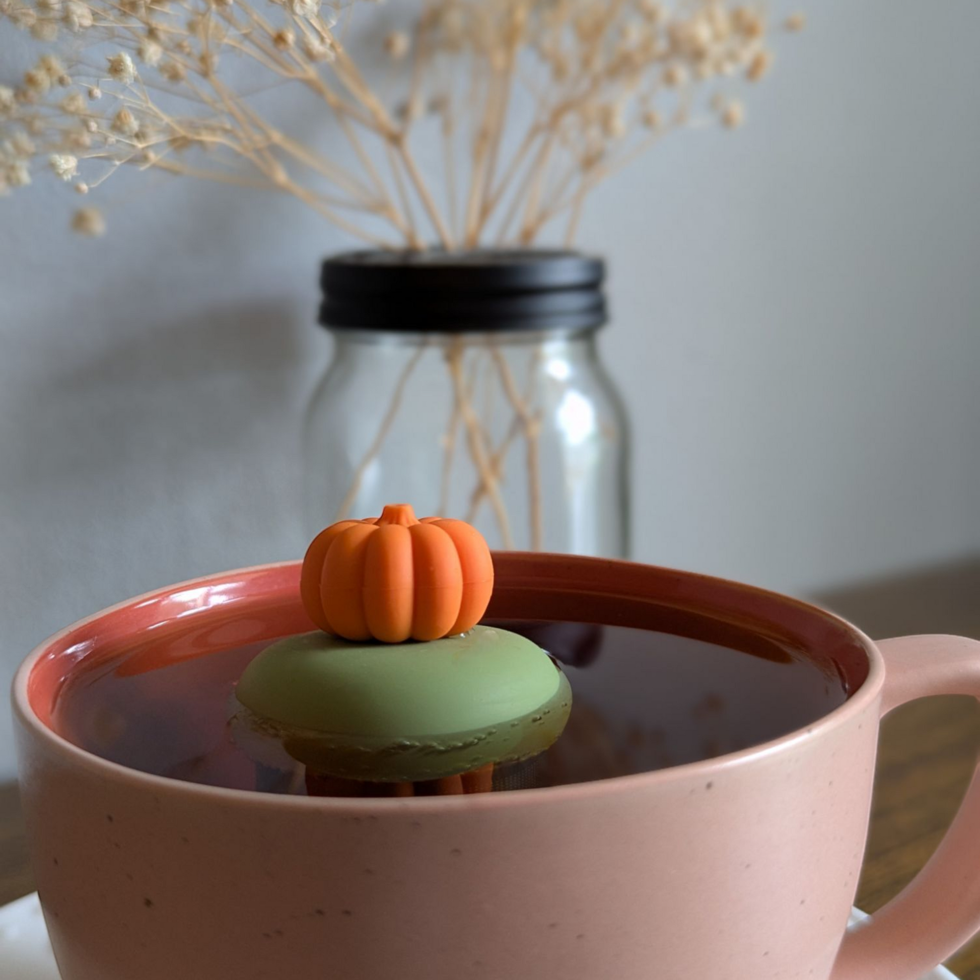 3D orange pumpkin tea infuser for loose leaf tea with a fine mesh stainless steel tea strainer that reads FLOAT2INFUSE on a green base  floating in a pink tea mug with flowers in the background