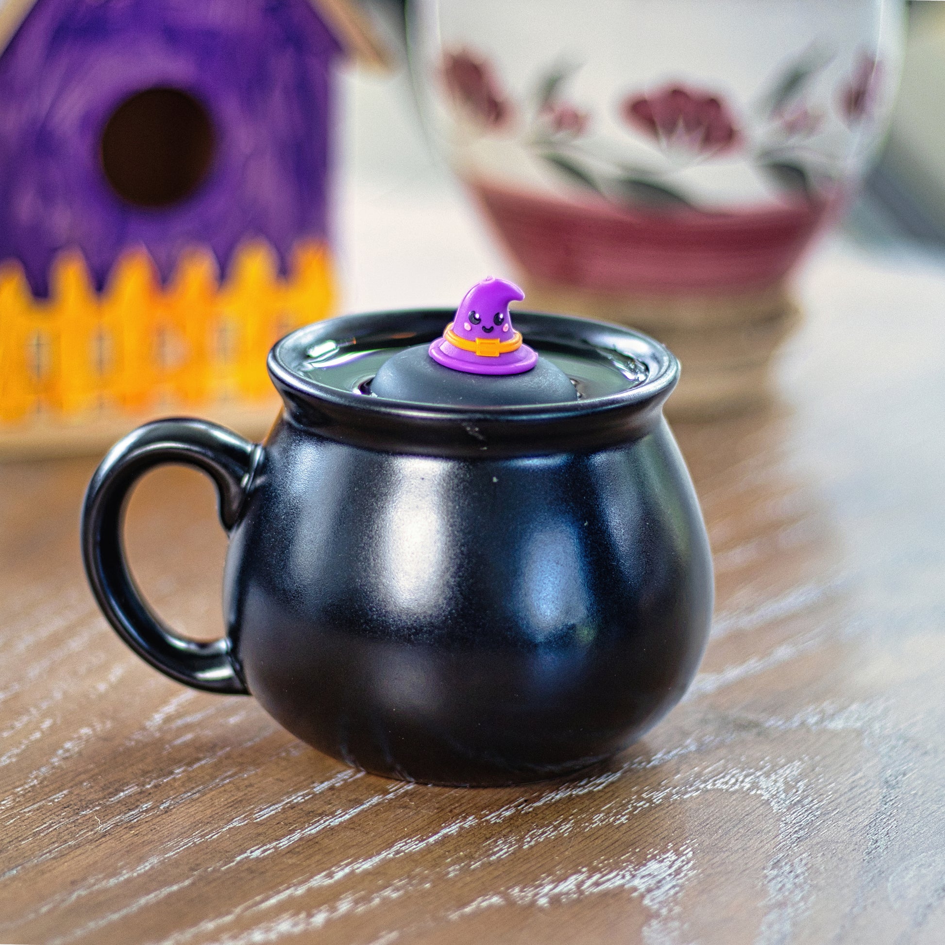 purple witch hat with a smiling face and an orange buckle tea infuser for loose leaf tea with a fine mesh stainless steel tea strainer that reads FLOAT2INFUSE on a black base floating in a black cauldron shaped tea mug with a bird house and a flower vase in the background
