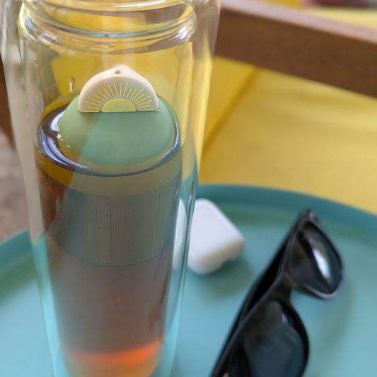 yellow sunrise tea infuser for loose leaf tea with a fine mesh stainless steel tea strainer that reads FLOAT2INFUSE on a green base floating in a tall glass travel tea mug with sunglasses and patio furniture in the background