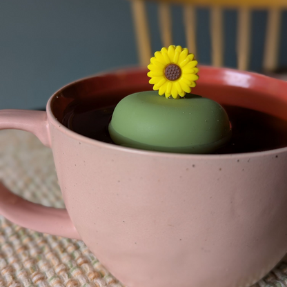 yellow sunflower tea infuser for loose leaf tea with a fine mesh stainless steel tea strainer that reads FLOAT2INFUSE on a green base  floating in a pink tea mug with a chair in the background