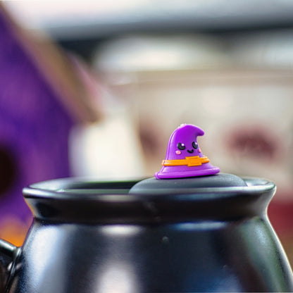 purple witch hat with a smiling face and an orange buckle tea infuser for loose leaf tea with a fine mesh stainless steel tea strainer that reads FLOAT2INFUSE on a black base floating in a black cauldron shaped tea mug 