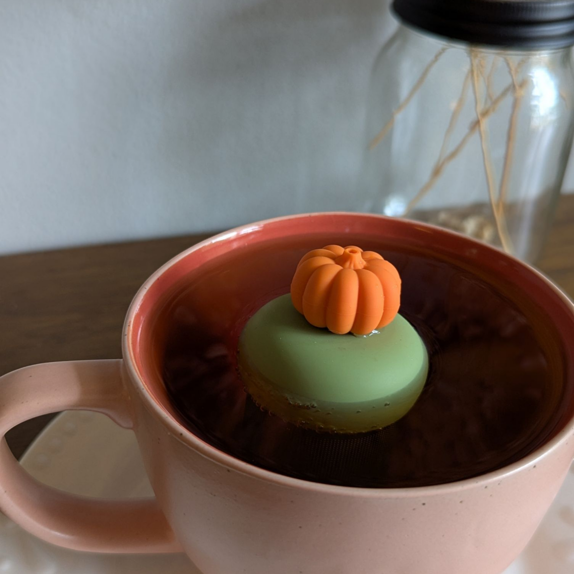 3D orange pumpkin tea infuser for loose leaf tea with a fine mesh stainless steel tea strainer that reads FLOAT2INFUSE on a green base  floating in a pink tea mug with flowers in the background