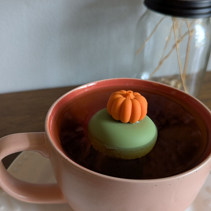 3D orange pumpkin tea infuser for loose leaf tea with a fine mesh stainless steel tea strainer that reads FLOAT2INFUSE on a green base  floating in a pink tea mug with flowers in the background