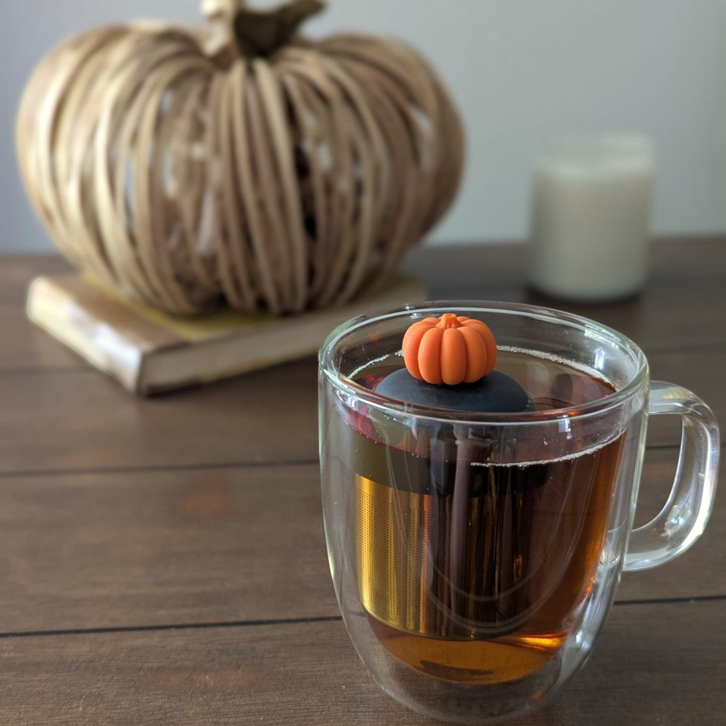 orange pumpkin tea infuser for loose leaf tea  with a fine mesh stainless steel strainer that reads FLOAT2INFUSE on a black base floating in a glass tea mug with a decorative pumpkin and a candle in the  background