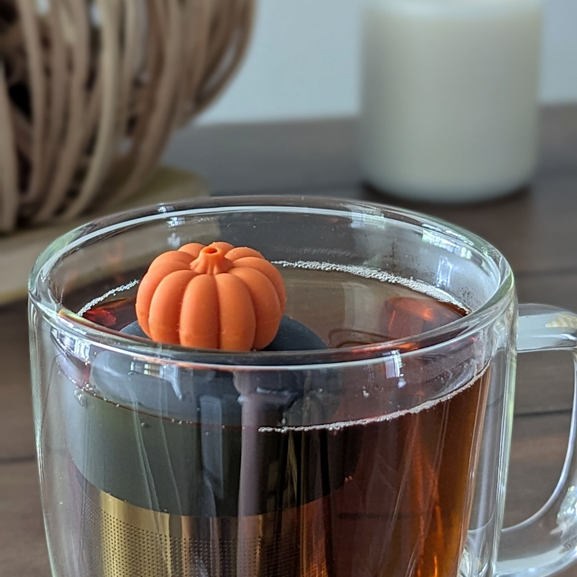 orange pumpkin tea infuser for loose leaf tea  with a fine mesh stainless steel strainer that reads FLOAT2INFUSE on a black base floating in a glass tea mug with a decorative pumpkin and a candle in the background