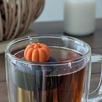 orange pumpkin tea infuser for loose leaf tea  with a fine mesh stainless steel strainer that reads FLOAT2INFUSE on a black base floating in a glass tea mug with a decorative pumpkin and a candle in the background