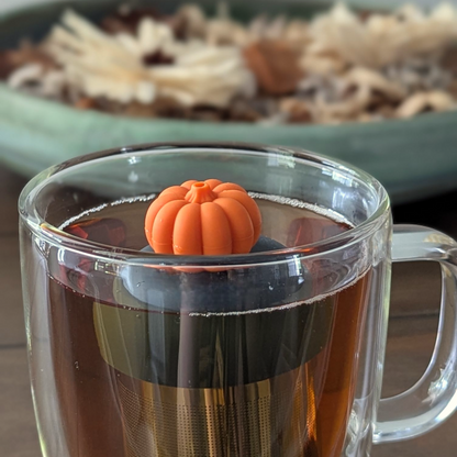 orange pumpkin tea infuser for loose leaf tea  with a fine mesh stainless steel strainer that reads FLOAT2INFUSE on a black base floating in a glass tea mug with dried herbs in the  background