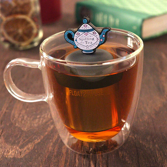 blue and white tea kettle that reads "spilling the tea"  tea infuser for loose leaf tea with a fine mesh stainless steel tea infuser that reads FLOAT2INFUSE on a black base  floating in a glass tea mug with  loose leaf tea and books in the background